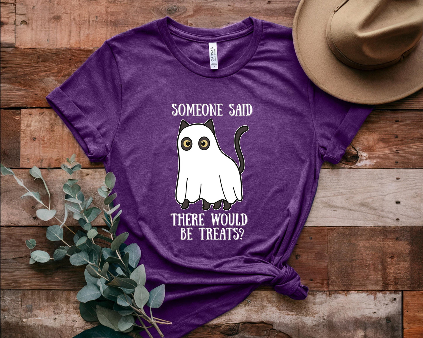 Purple t-shirt with a ghost graphic and text on a wooden surface with a hat and plant.