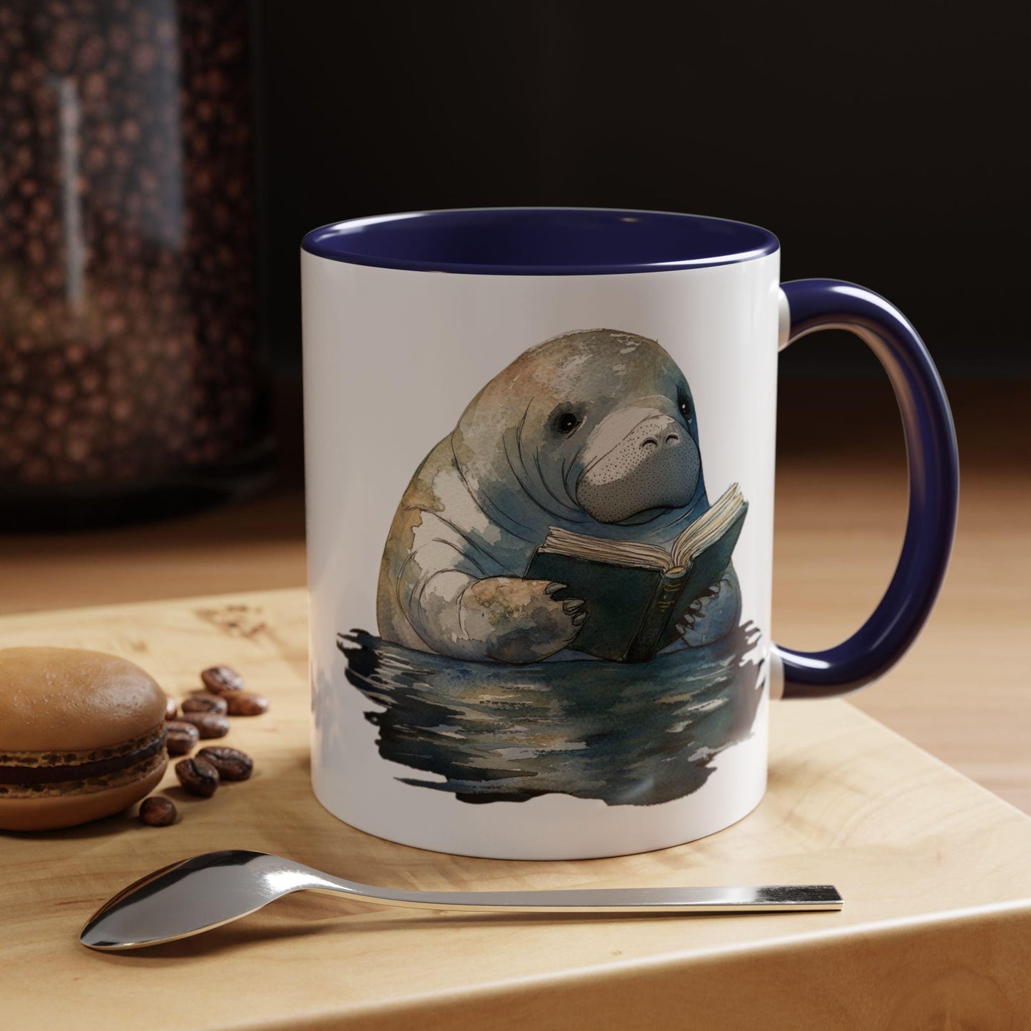 Mug with a manatee reading a book design on a table with coffee beans and a spoon.