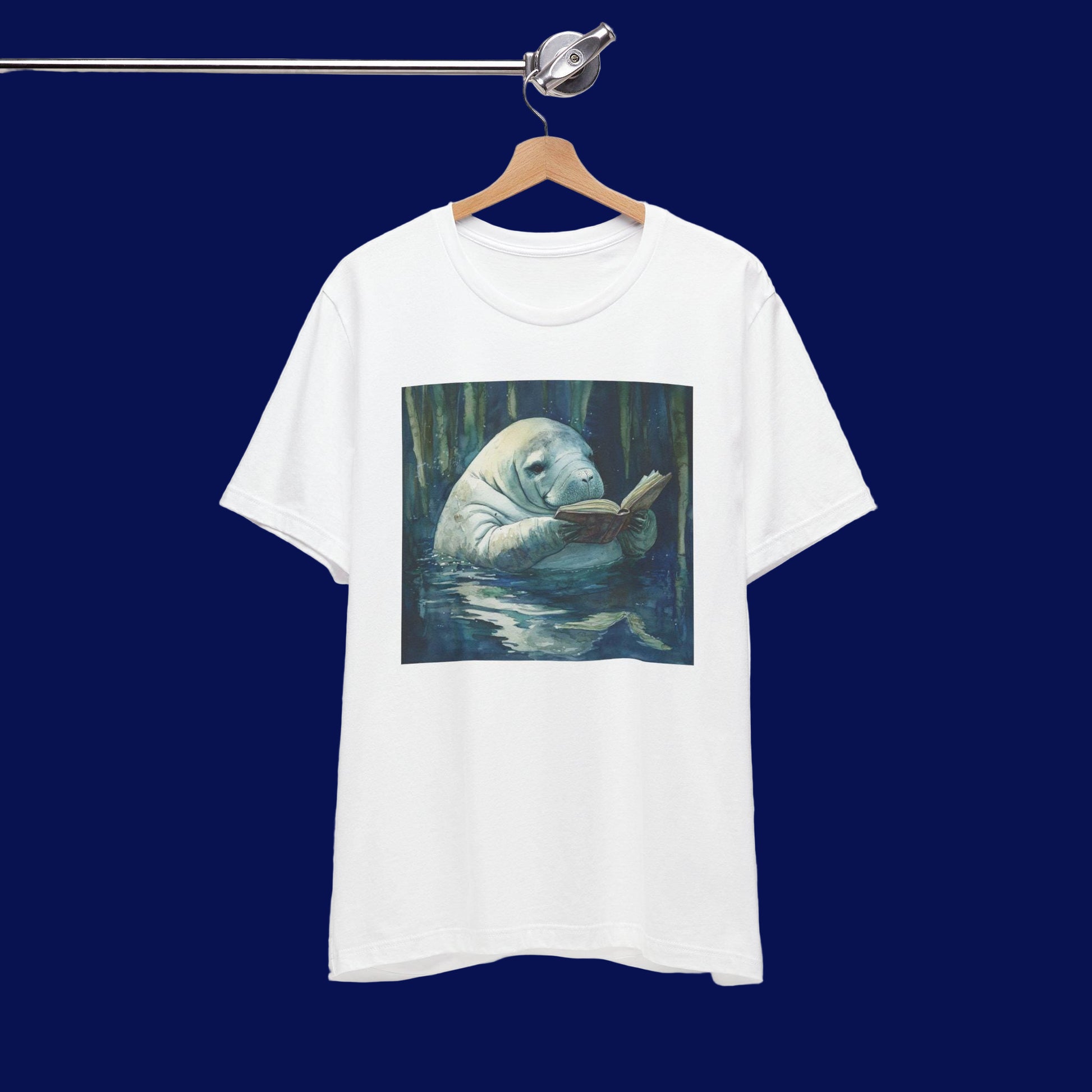 White t-shirt with a graphic of a manatee reading a book in water, hanging on a wooden hanger against a white background.