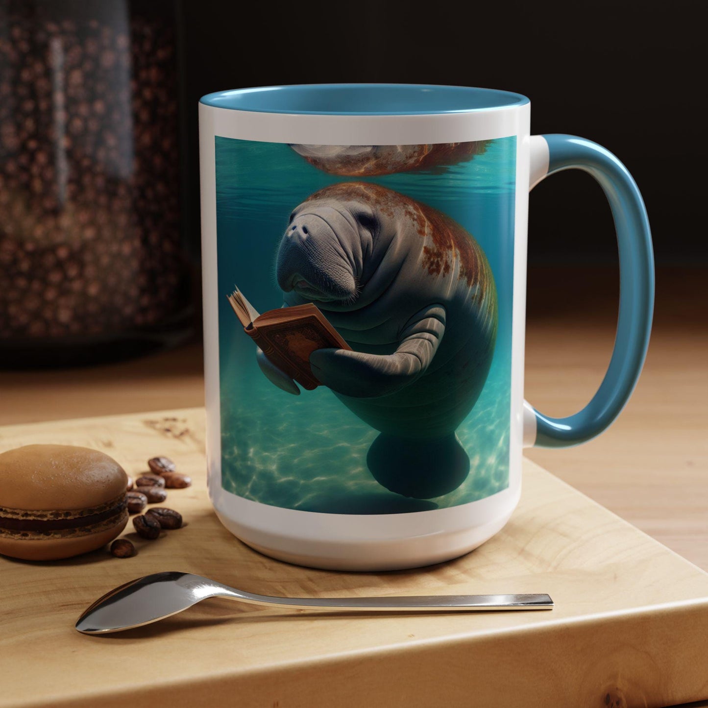 Mug with a manatee reading a book design on a table with coffee beans and a macaron.