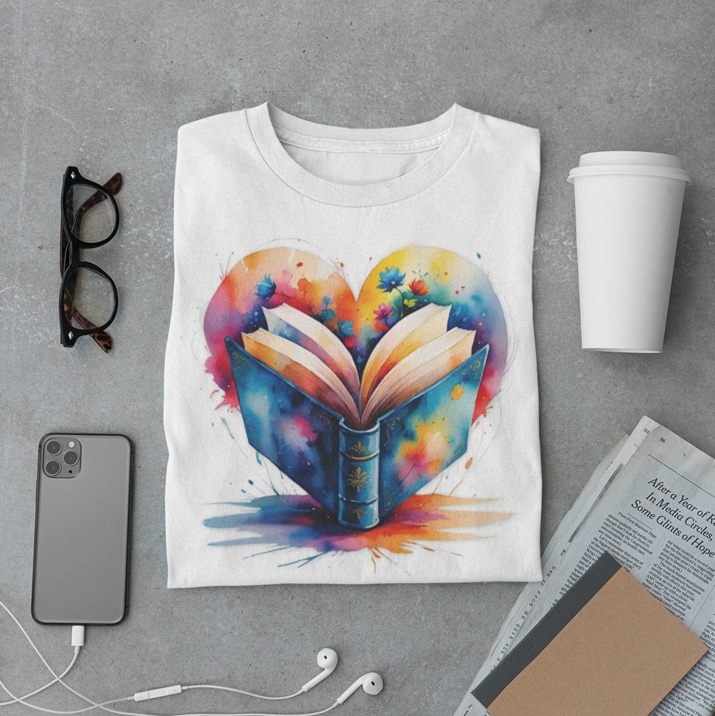 White t-shirt with colorful heart and book design on a gray surface with phone, glasses, and cup.