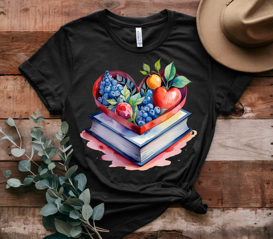 Black t-shirt with a colorful design of books and heart-shaped fruits on a wooden surface.