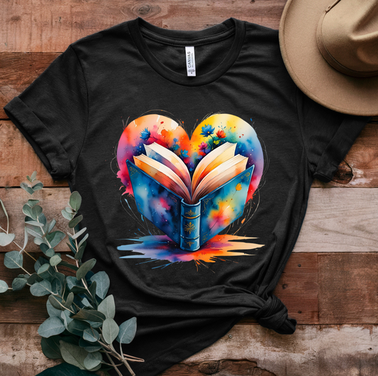 Black t-shirt with a colorful heart-shaped book design on a wooden surface with a hat and plant.