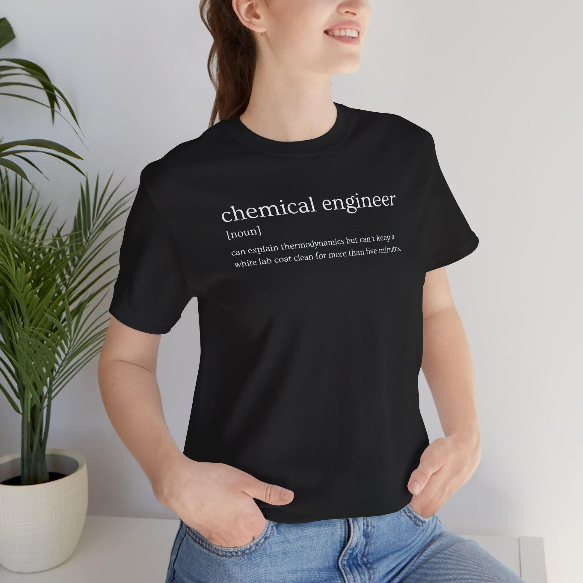 Person wearing a black t-shirt with a humorous definition of a chemical engineer, standing indoors next to a plant.