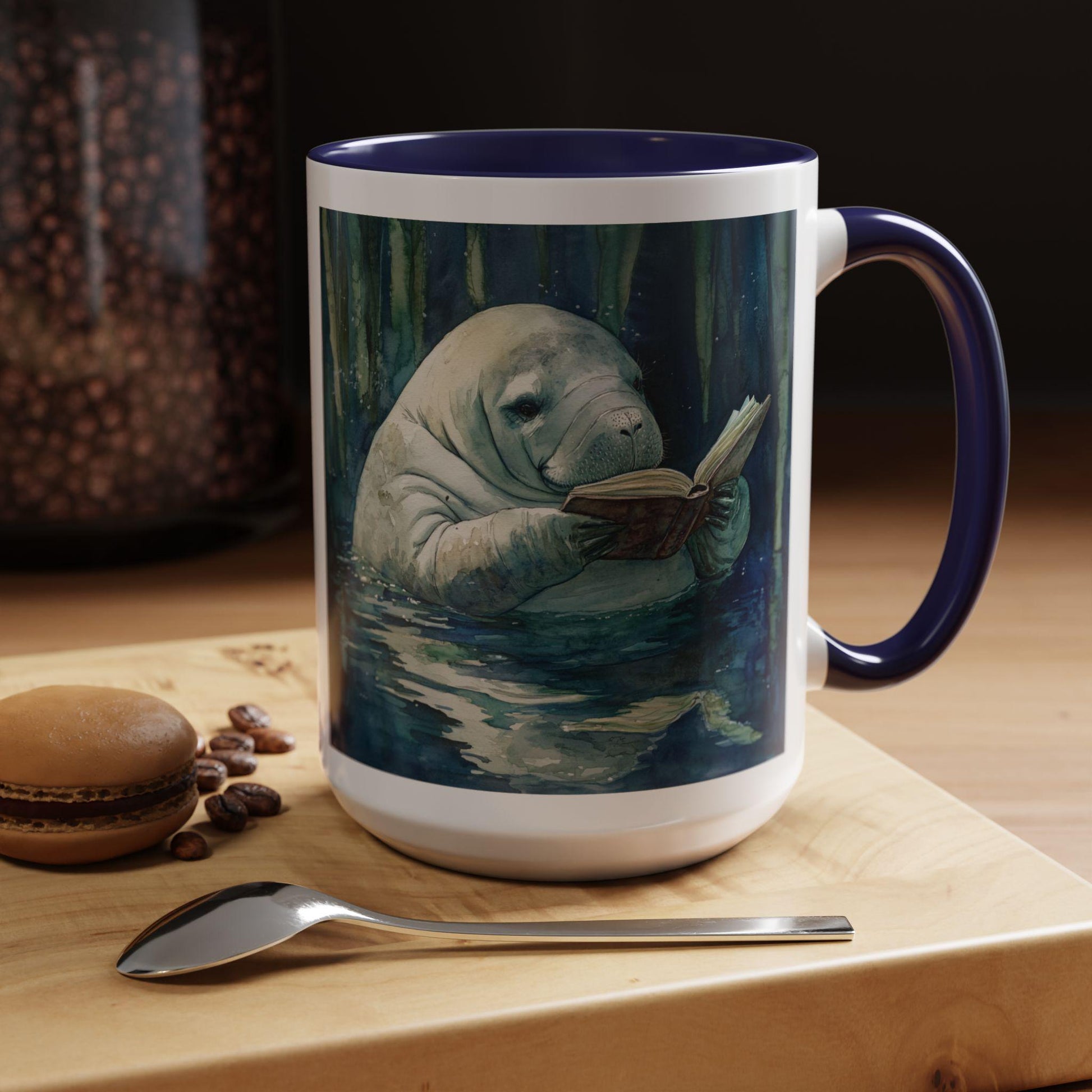 Mug with a design of a manatee reading a book on a wooden surface with coffee beans and a macaron.