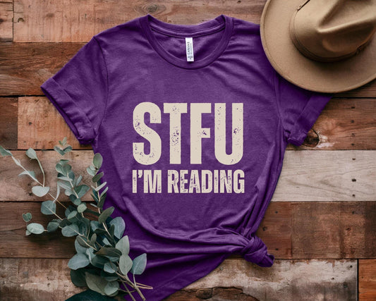 Purple t-shirt with 'STFU I'm Reading' text on a wooden surface with a hat and plant.