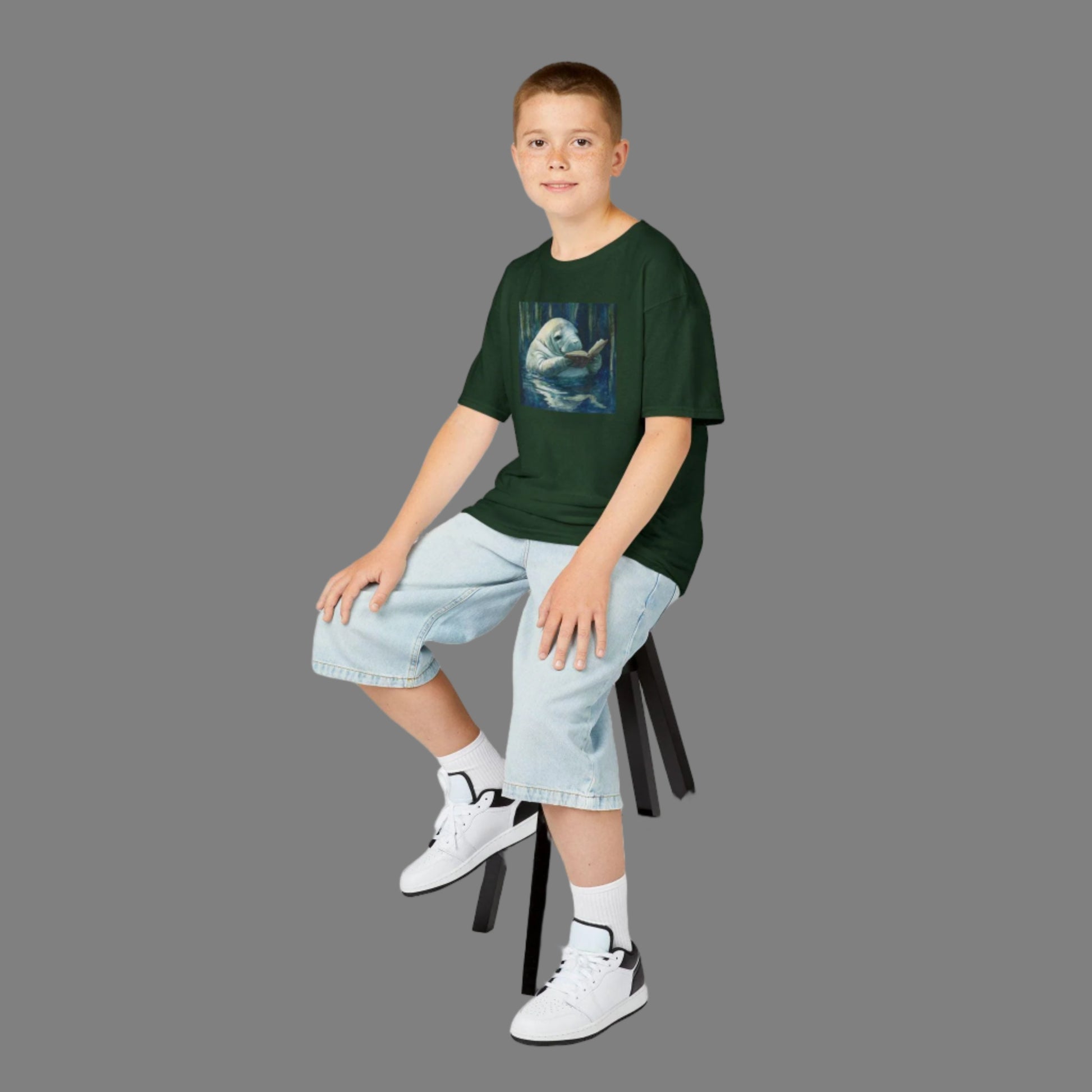 Young boy sitting on a stool wearing a green t-shirt with a graphic design, light blue jeans, and white sneakers.