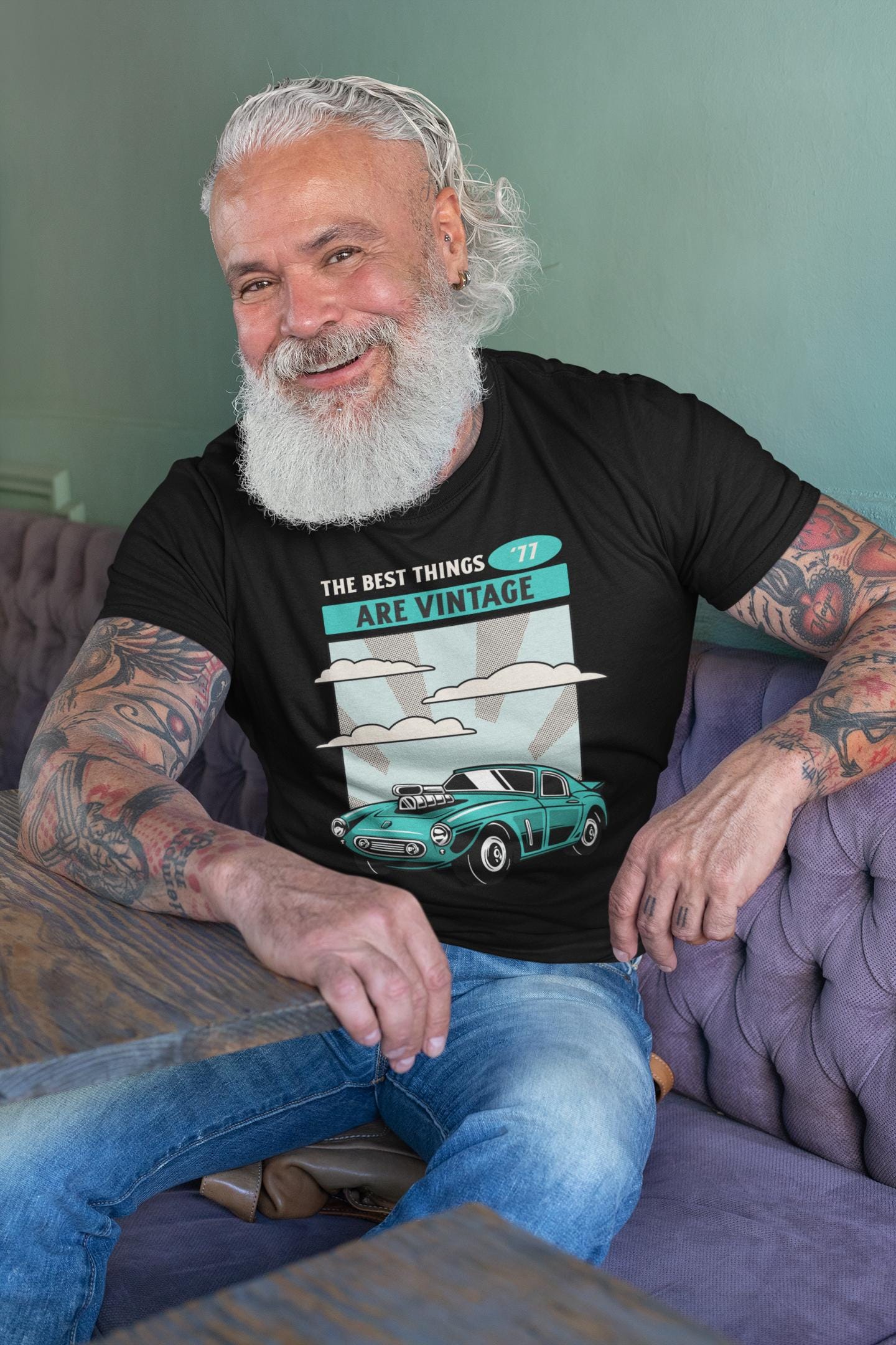Man with tattoos and a beard wearing a black t-shirt with a graphic design, sitting on a purple couch.