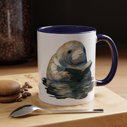 Mug with a manatee reading a book design on a table with coffee beans and a spoon.