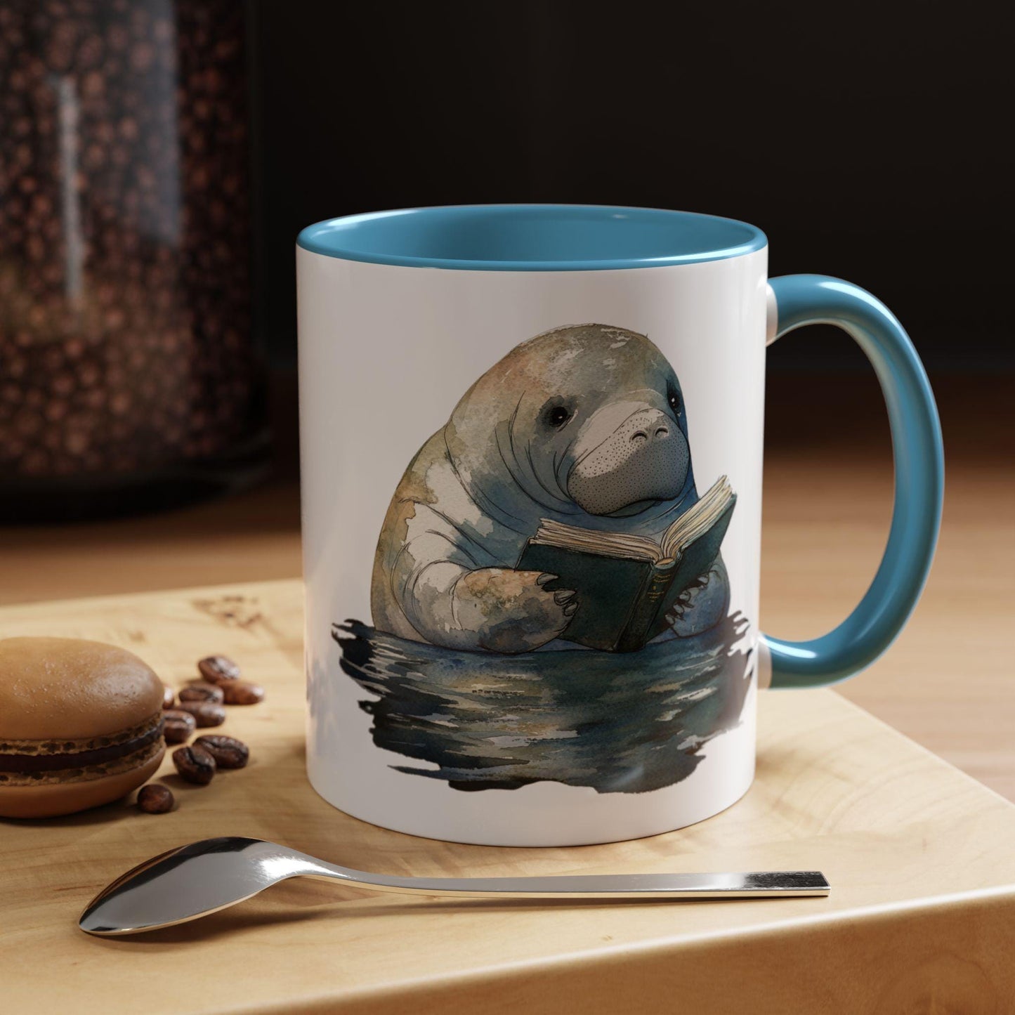Mug with a manatee reading a book design on a table with coffee beans and a spoon.