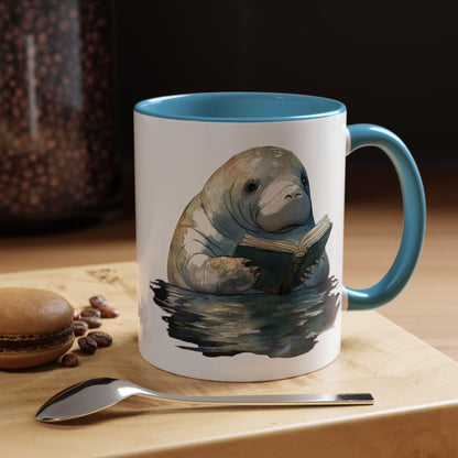 Mug with a manatee reading a book design on a table with coffee beans and a spoon.
