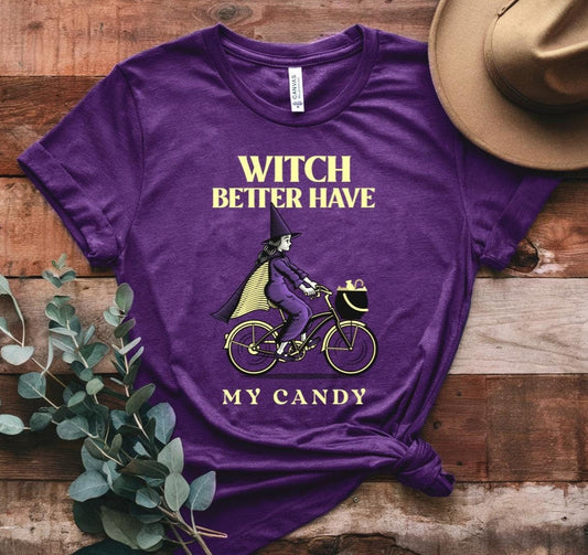 Purple t-shirt with Halloween-themed text and graphics on a wooden surface with a hat and eucalyptus leaves.