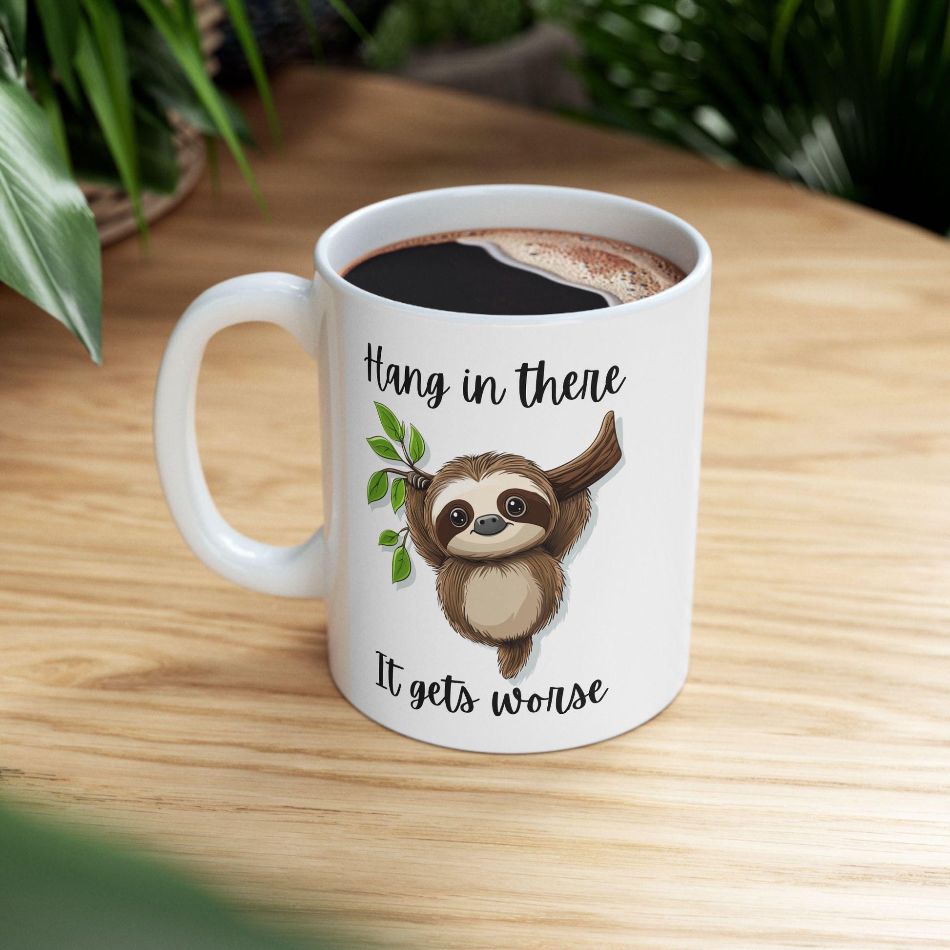 White mug with sloth design and text 'Hang in there It gets worse' on a wooden surface.